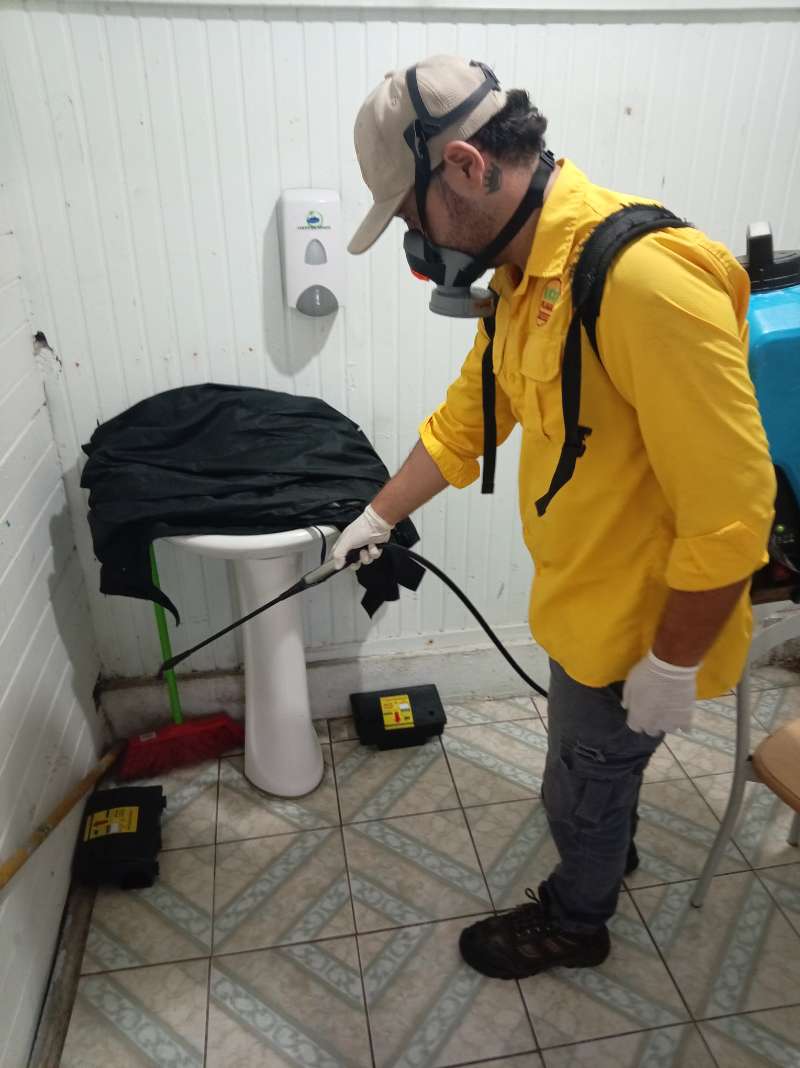 Técnico de EcoPlagas con respirador, gorra y guantes aplicando tratamiento en un baño comercial